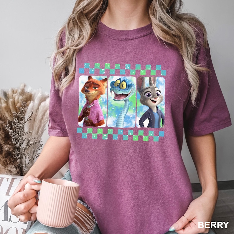 May include: Berry-colored t-shirt featuring three cartoon characters: a fox in a pink shirt, a snake, and a rabbit in a blue jacket. The characters are set against a colorful background and framed by a checkered pattern. The word "BERRY" is printed at the bottom.