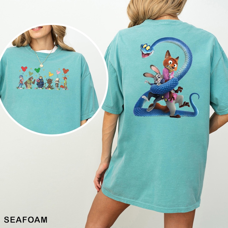 Op de afbeelding: Zeegroen t-shirt met cartoonafbeeldingen op de voor- en achterkant. De afbeelding op de voorkant toont stripfiguren met hartvormige ballonnen. De afbeelding op de achterkant toont een vos en een konijn die door een blauwe slang worden omarmd.