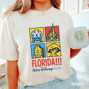 Może przedstawiać: Kremowa koszulka z kolorową grafiką czterech kwadrat&oacute;w z ikonami Disney World. Słowo "FLORIDA!!!" jest wydrukowane na czerwono, a pod nim "Walt Disney World" na niebiesko. Na zdjęciu trzymany jest biały kubek z uśmiechniętą buzią.