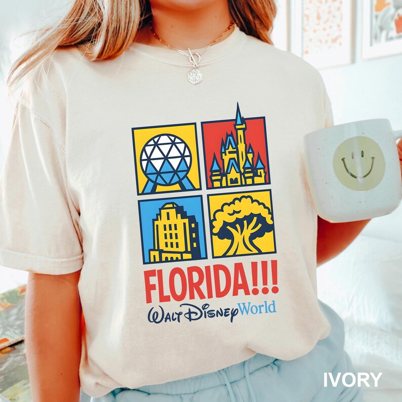 Może przedstawiać: Kremowa koszulka z kolorową grafiką czterech kwadrat&oacute;w z ikonami Disney World. Słowo "FLORIDA!!!" jest wydrukowane na czerwono, a pod nim "Walt Disney World" na niebiesko. Na zdjęciu trzymany jest biały kubek z uśmiechniętą buzią.
