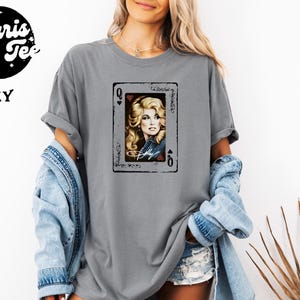 May include: A grey short-sleeve t-shirt featuring a graphic of a playing card with a portrait of a blonde woman and the text "Dolly" in white script. The card design includes the letter Q and a heart symbol in the corners, with a distressed border.