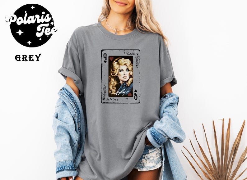 May include: A grey short-sleeve t-shirt featuring a graphic of a playing card with a portrait of a blonde woman and the text "Dolly" in white script. The card design includes the letter Q and a heart symbol in the corners, with a distressed border.
