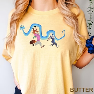 Op de afbeelding: Geel T-shirt met een cartoon illustratie van een vos en een konijn die een blauwe slang vasthouden. De vos draagt een roze shirt en het konijn draagt een pak. Het woord "BUTTER" staat onderaan gedrukt.