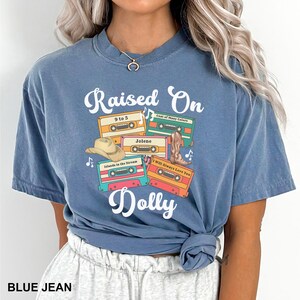 May include: A blue-jean colored short-sleeve t-shirt featuring a graphic print with the text Raised On Dolly. The design includes four colorful cassette tapes labeled 9 to 5, Coat of Many Colors, Jolene, Islands in the Stream, and I Will Always Love You, alongside a cowboy hat and a boot.