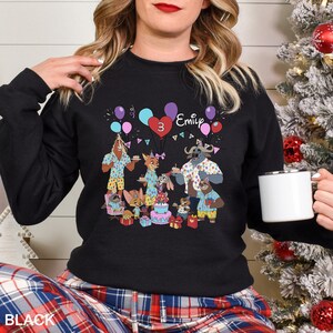 May include: Black crewneck sweatshirt featuring a colorful cartoon birthday party scene with animal characters, balloons, a cake, and the name "Emily" with the number "3". The word "BLACK" is printed at the bottom.