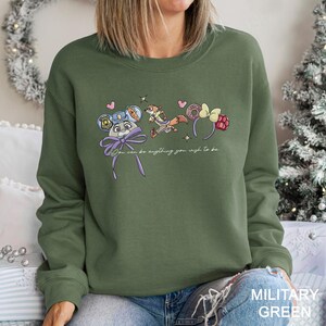 May include: A military green sweatshirt with a playful design. The design features cartoon characters and accessories, including a mouse with a police hat, a fox, and headbands. The text "You can be anything you wish to be" is also included.