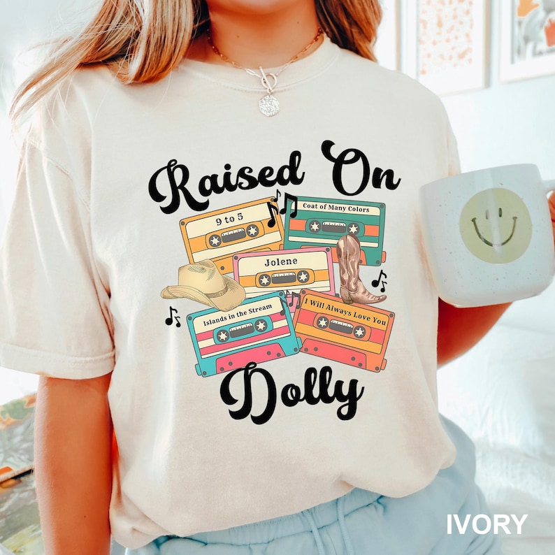 May include: An ivory t-shirt featuring a graphic print with the text "Raised On Dolly" and four colorful cassette tapes labeled "9 to 5," "Coat of Many Colors," "Jolene," "Islands in the Stream," and "I Will Always Love You," plus a cowboy hat and boot.