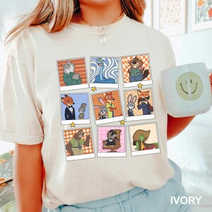 May include: Ivory-colored t-shirt featuring a grid of cartoon character portraits in polaroid frames. The portraits depict various animal characters in different outfits and poses. A white mug with a smiley face is also visible.