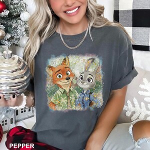 May include: Dark gray t-shirt featuring a colorful graphic of Nick Wilde and Judy Hopps from Zootopia. Nick is wearing a Hawaiian shirt, and Judy is in a police uniform. The shirt has a relaxed fit and rolled sleeves.