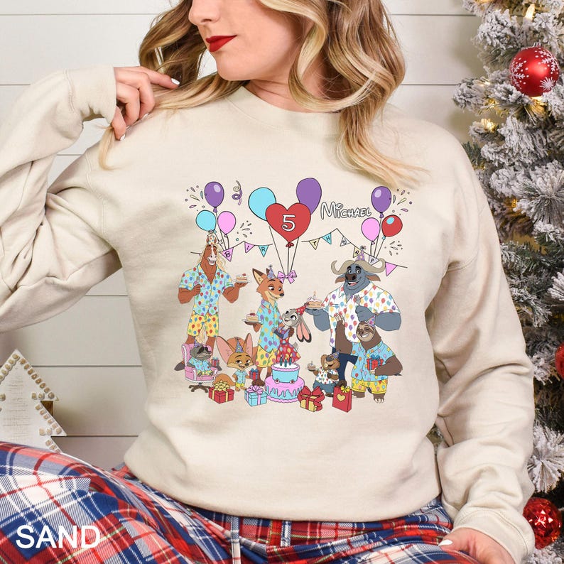 May include: A sand-colored sweatshirt featuring a colorful cartoon illustration of a birthday party with animal characters. The design includes balloons, a heart-shaped balloon with the number 5, and the name "Michael".