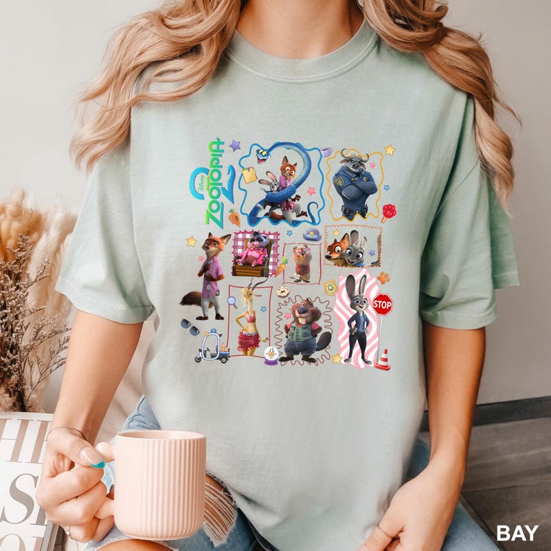May include: A light green t-shirt featuring a graphic print of various Zootopia characters including Judy Hopps and Nick Wilde. The design includes multiple framed scenes, the Zootopia logo, and small icons like a stop sign, sunglasses, and a scooter.