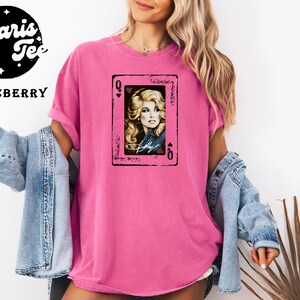 May include: A bright pink t-shirt featuring a central graphic of a playing card with the letter Q in the corners. The card displays a portrait of a blonde woman with a signature below the image. The shirt is worn with a light blue denim jacket.