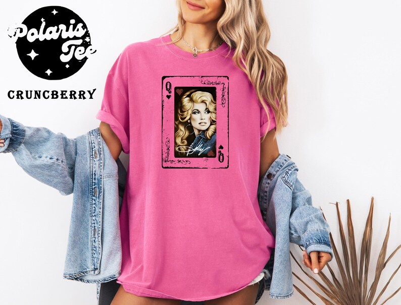 May include: A bright pink t-shirt featuring a central graphic of a playing card with the letter Q in the corners. The card displays a portrait of a blonde woman with a signature below the image. The shirt is worn with a light blue denim jacket.