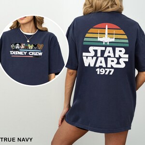 May include: Navy blue t-shirt with a Disney Crew graphic on the front and a Star Wars design on the back. The Star Wars design features a retro sunset, a spaceship, and the text "STAR WARS 1977".