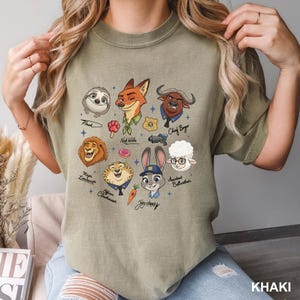 K&ouml;nnte beinhalten: Khaki-farbenes T-Shirt mit Cartoon-Illustrationen von Charakteren aus dem Animationsfilm "Zoomania". Das Design umfasst Flash, Nick Wilde, Judy Hopps, Chief Bogo und andere. Das Wort "KHAKI" ist unten aufgedruckt.
