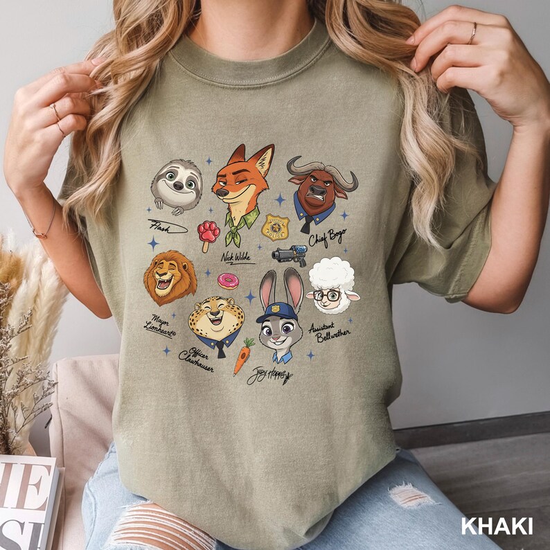 K&ouml;nnte beinhalten: Khaki-farbenes T-Shirt mit Cartoon-Illustrationen von Charakteren aus dem Animationsfilm "Zoomania". Das Design umfasst Flash, Nick Wilde, Judy Hopps, Chief Bogo und andere. Das Wort "KHAKI" ist unten aufgedruckt.