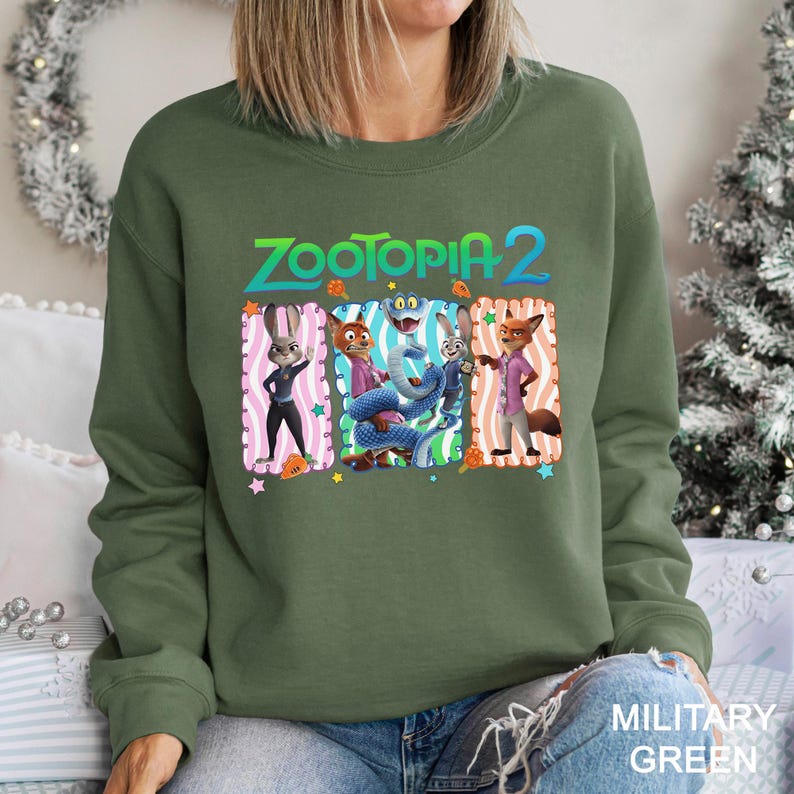 May include: Military green sweatshirt featuring the text "Zootopia 2" above a graphic of characters from the animated film. The graphic includes a bunny, fox, and other characters in colorful frames. The sweatshirt is a crew neck style.
