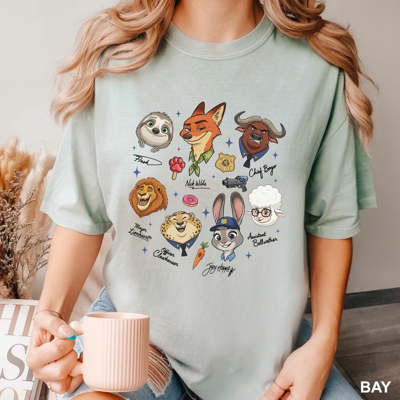 K&ouml;nnte beinhalten: Hellgr&uuml;nes T-Shirt mit Cartoon-Illustrationen von Zootopia-Charakteren, darunter Flash, Nick Wilde, Chief Bogo und Judy Hopps. Auf dem T-Shirt steht das Wort "BAY" unten rechts.