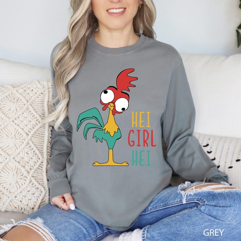 May include: A grey long-sleeve shirt featuring a cartoon rooster with red, yellow, and teal accents. The shirt has the text "HEI GIRL HEI" in colorful letters. The word "GREY" is printed at the bottom right.