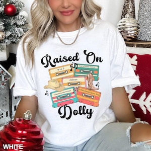 May include: A white t-shirt featuring a graphic print with the text "Raised On Dolly" in black script. The design includes four colorful cassette tapes labeled "9 to 5," "Coat of Many Colors," "Jolene," "Islands in the Stream," and "I Will Always Love You," along with a tan cowboy hat and a brown cowboy boot.