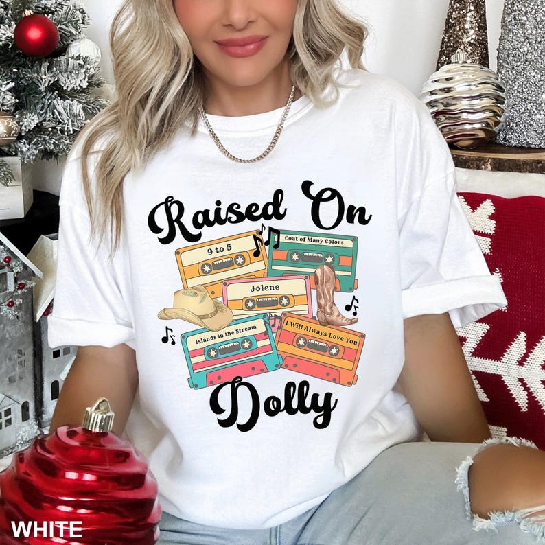 May include: A white t-shirt featuring a graphic print with the text "Raised On Dolly" in black script. The design includes four colorful cassette tapes labeled "9 to 5," "Coat of Many Colors," "Jolene," "Islands in the Stream," and "I Will Always Love You," along with a tan cowboy hat and a brown cowboy boot.