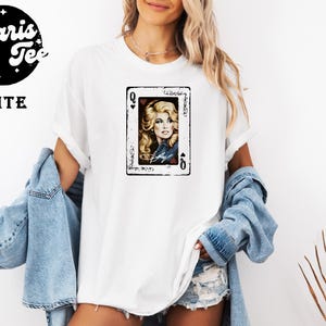 May include: A white short-sleeved t-shirt featuring a graphic print of a playing card with the letter Q and a portrait of a blonde woman. The text Polaris Tee and WHITE are printed in the upper left corner. The shirt is worn with a blue denim jacket.