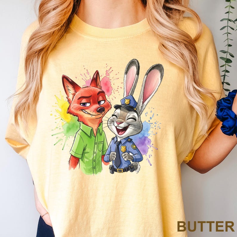 May include: Yellow t-shirt featuring a colorful graphic of Nick Wilde the fox and Judy Hopps the rabbit from Zootopia. Nick is wearing a green shirt, and Judy is in a police uniform. The word "BUTTER" is printed at the bottom.