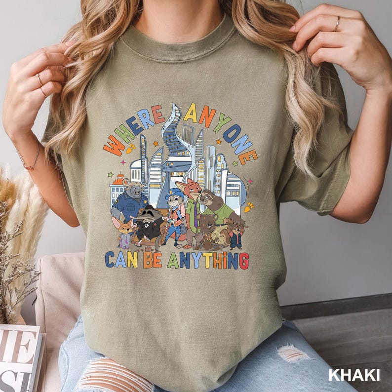 May include: Khaki-colored t-shirt featuring a colorful graphic with the text "WHERE ANYONE CAN BE ANYTHING." The graphic includes cartoon characters and a cityscape. The shirt is a casual style.