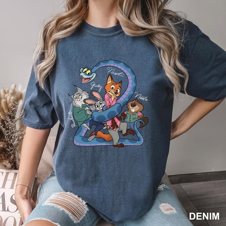 Peut inclure: T-shirt bleu denim avec un dessin anim&eacute; de personnages de film. Le dessin comprend un serpent et plusieurs personnages d'animaux, avec des noms &eacute;crits au-dessus. Le mot "DENIM" est imprim&eacute; en bas &agrave; droite.