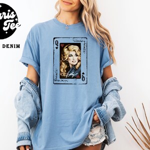 May include: A light blue t-shirt featuring a graphic print of a playing card with the letter Q in the corners. The card displays a portrait of a blonde person with long hair, wearing a denim jacket, with the name Dolly written in white script.