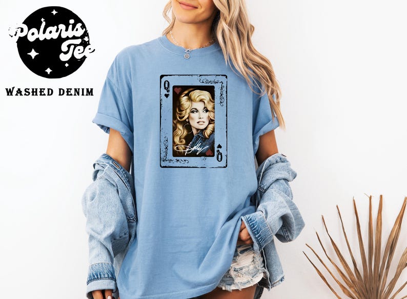 May include: A light blue t-shirt featuring a graphic print of a playing card with the letter Q in the corners. The card displays a portrait of a blonde person with long hair, wearing a denim jacket, with the name Dolly written in white script.