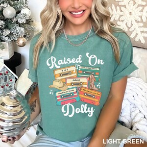 May include: A light green t-shirt featuring the text Raised On Dolly in white script. The graphic includes four colorful cassette tapes labeled 9 to 5, Coat of Many Colors, Jolene, and Islands in the Stream, along with a cowboy hat and boot.