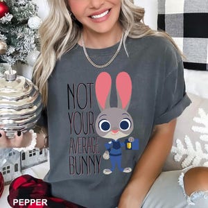 May include: Dark gray t-shirt featuring a cartoon bunny character with pink ears and a blue uniform, holding a blue briefcase. The text "NOT YOUR AVERAGE BUNNY" is printed on the shirt in black and pink.