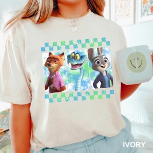 May include: Ivory-colored t-shirt featuring a graphic of three animated characters: a fox, a snake, and a rabbit. The design is framed with a blue and green checkered pattern. A white mug with a smiley face is held in the image.