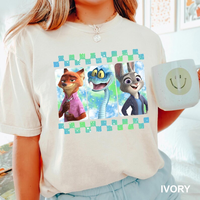 May include: Ivory-colored t-shirt featuring a graphic of three animated characters: a fox, a snake, and a rabbit. The design is framed with a blue and green checkered pattern. A white mug with a smiley face is held in the image.