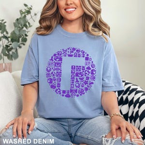 May include: A light blue t-shirt with a large purple graphic of the letter "G" surrounded by various video game icons. The shirt is made of a soft material and has the words "WASHED DENIM" printed at the bottom.