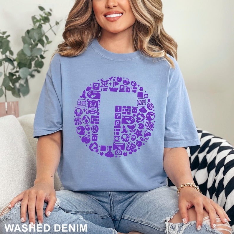 May include: A light blue t-shirt with a large purple graphic of the letter "G" surrounded by various video game icons. The shirt is made of a soft material and has the words "WASHED DENIM" printed at the bottom.