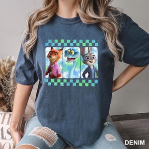 May include: A denim blue t-shirt featuring a graphic of three animated characters: a fox in a pink shirt, a snake, and a rabbit in a blue jacket. The graphic is framed with a checkered pattern in green and blue.