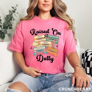 May include: A bright pink t-shirt featuring a graphic print with the text "Raised On Dolly" in black script. The design includes four colorful cassette tapes labeled with song titles, a tan cowboy hat, a brown cowboy boot, and musical notes.