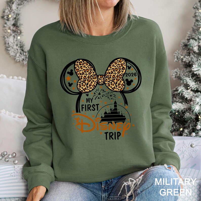 May include: Military green sweatshirt with a graphic design featuring Mickey Mouse ears, a leopard print bow, and the text "My First Disney Trip" in orange. The year "2026" is also included in the design.