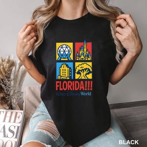 Może przedstawiać: Czarny t-shirt z kolorową grafiką przedstawiającą zabytki Florydy, w tym kulę, zamek, budynek i drzewo. Słowo "FLORIDA!!!" jest w kolorze czerwonym, z napisem "Walt Disney World" poniżej. Okrągły dekolt.