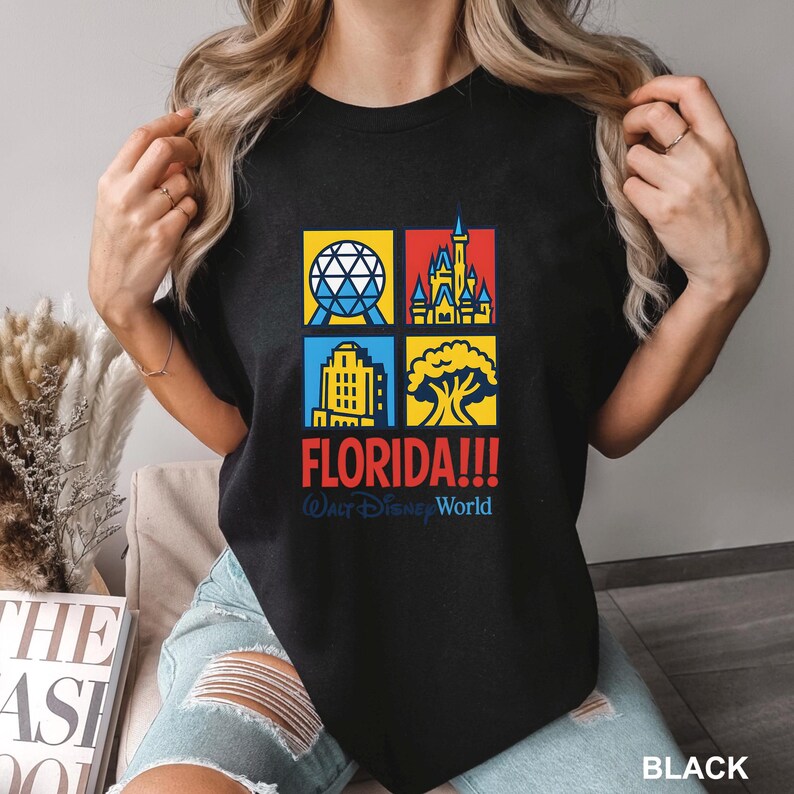 Może przedstawiać: Czarny t-shirt z kolorową grafiką przedstawiającą zabytki Florydy, w tym kulę, zamek, budynek i drzewo. Słowo "FLORIDA!!!" jest w kolorze czerwonym, z napisem "Walt Disney World" poniżej. Okrągły dekolt.