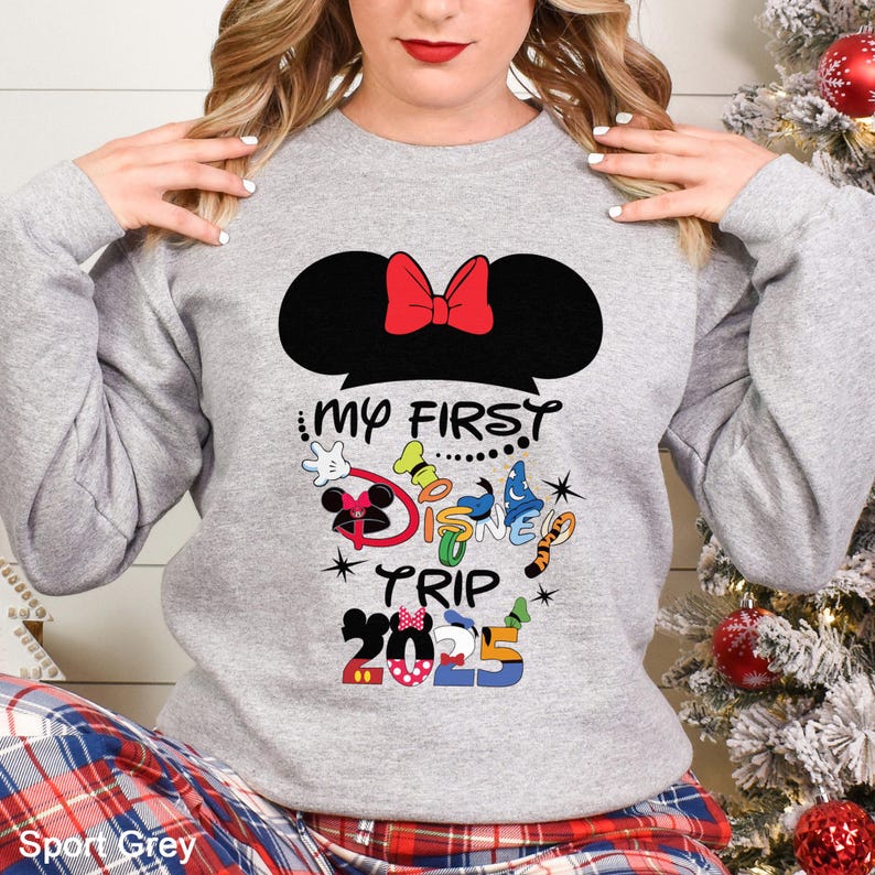 Puede incluir: Una sudadera gris claro con una silueta negra de Minnie Mouse y un lazo rojo. El texto "MY FIRST DISNEY TRIP 2025" est&aacute; impreso debajo, junto con gr&aacute;ficos coloridos con tem&aacute;tica de Disney. La sudadera es de material suave.