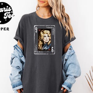 May include: A charcoal gray short-sleeve t-shirt featuring a graphic of a playing card with a portrait of a blonde person and the text "Dolly". The shirt has a crew neckline and a relaxed fit. A logo in the corner reads "Polaris Tee PEPPER".