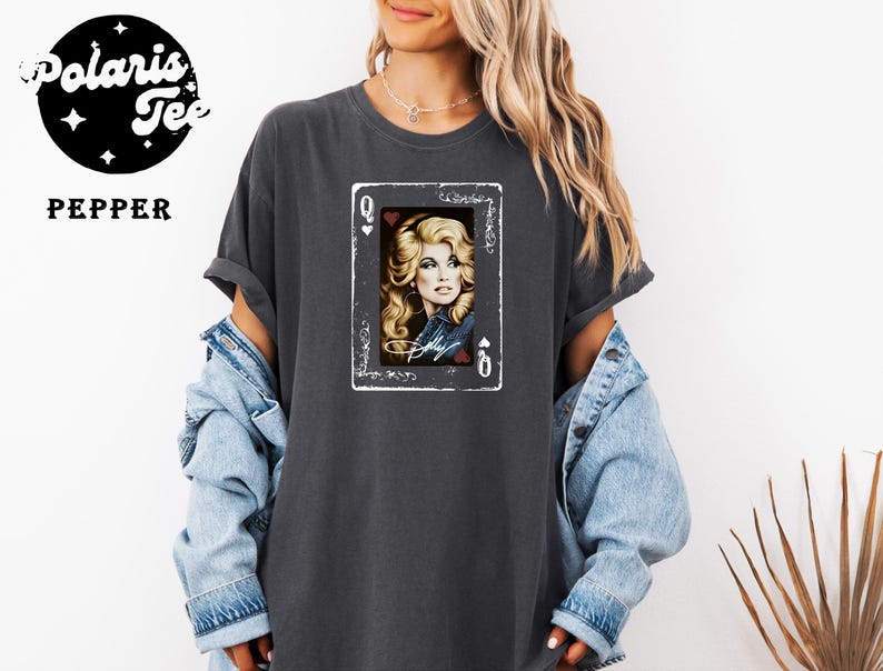 May include: A charcoal gray short-sleeve t-shirt featuring a graphic of a playing card with a portrait of a blonde person and the text "Dolly". The shirt has a crew neckline and a relaxed fit. A logo in the corner reads "Polaris Tee PEPPER".