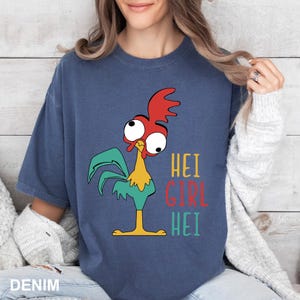 May include: Denim blue t-shirt featuring a cartoon rooster with red, green, and yellow accents. The text "HEI GIRL HEI" is printed in colorful letters on the right side of the rooster.