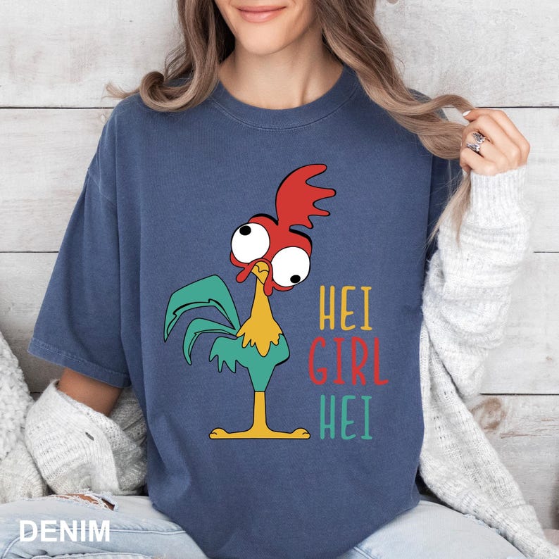 May include: Denim blue t-shirt featuring a cartoon rooster with red, green, and yellow accents. The text "HEI GIRL HEI" is printed in colorful letters on the right side of the rooster.