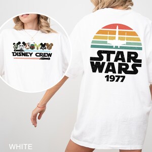 May include: White t-shirt with a "Disney Crew" design on the front, showcasing character illustrations and a lightsaber. The back features a "Star Wars 1977" graphic with a retro sunset and spaceship silhouette.