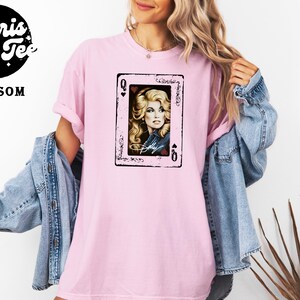 May include: A light pink short-sleeve t-shirt featuring a graphic print of a playing card with a portrait of a blonde woman and the text Dolly. The shirt is layered under a blue denim jacket. The Polaris Tee Blossom logo is in the top left corner.