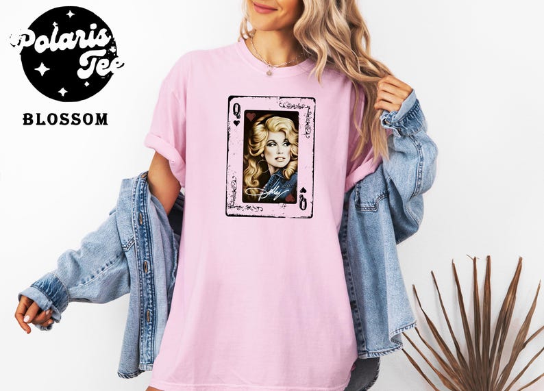 May include: A light pink short-sleeve t-shirt featuring a graphic print of a playing card with a portrait of a blonde woman and the text Dolly. The shirt is layered under a blue denim jacket. The Polaris Tee Blossom logo is in the top left corner.